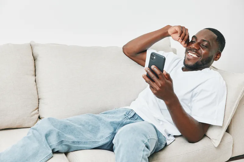 Smiling young Black man relaxing on a couch, laughing at his smartphone, wearing casual clothes, conveying happiness and joy in a bright indoor setting