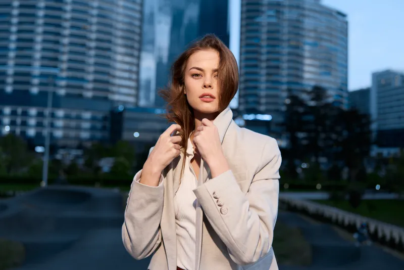 Confident woman in business attire, standing outdoor in urban setting with modern architecture, exuding professionalism and elegance during daytime.