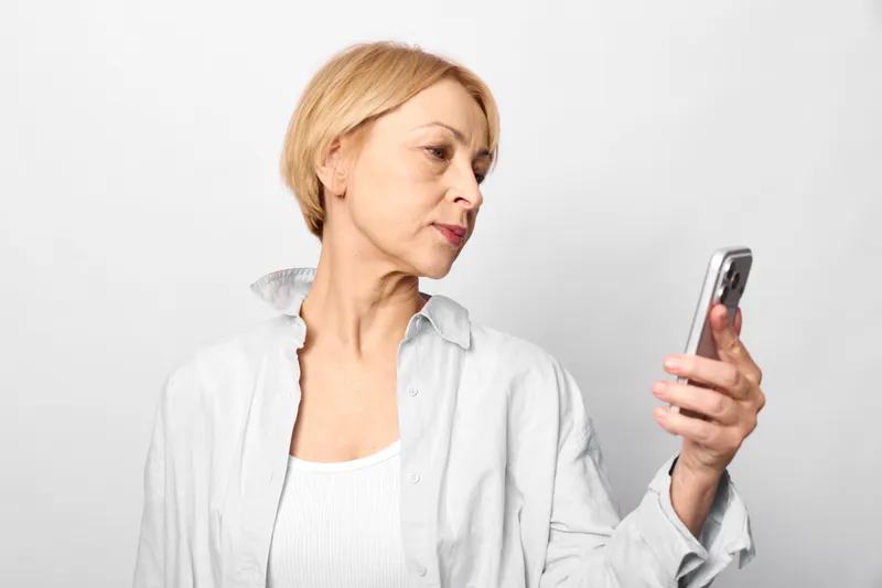 Senior woman shows stress and anxiety, staring at a smartphone with worry and tense expression against white wall, concept of mental health and loneliness.