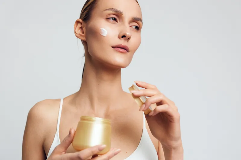 A woman applies a creamy moisturizer from a jar in a bright studio, clearly documenting a calm skincare routine. The scene emphasizes self care, facial hydration, and a focused beauty moment.