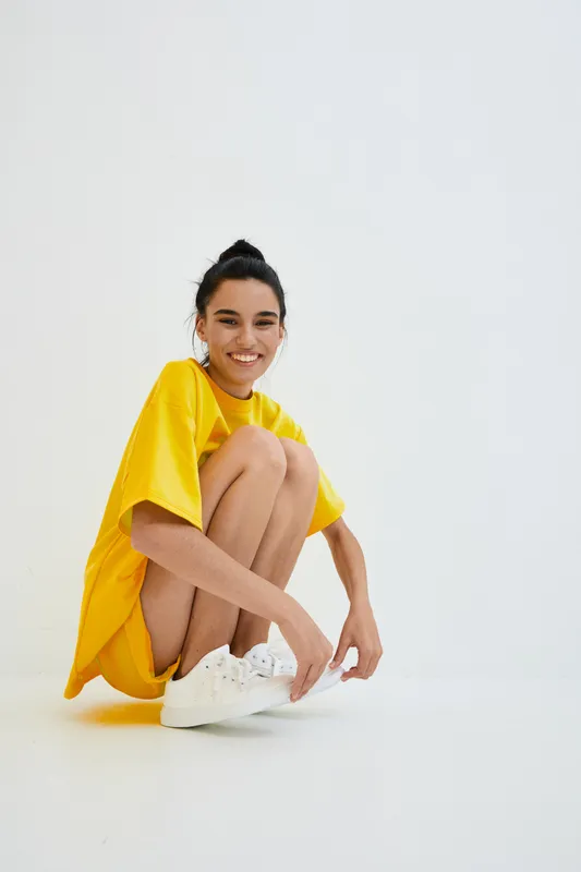 smiling woman in yellow oversized t-shirt and white sneakers sitting crouched on white seamless background, casual style, natural light, minimalistic mood