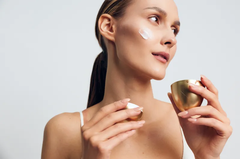 Young woman applying face cream on cheek with soft fingers holding golden jar. Close-up on white background. Concept of skincare, beauty routine, wellness, and self-care.