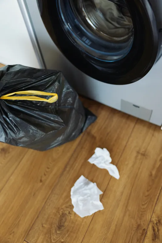 Washing machine and laundry scene with garbage and plastic bag on wooden floor, crumpled tissues beside a frontload washer door, household mess, dirty clothes and cleanup needed.