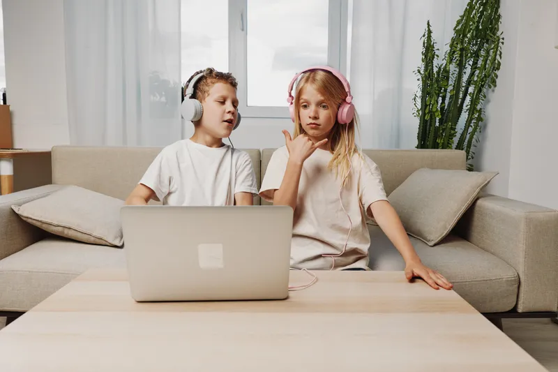 Two children with headphones sit on couch watching laptop, enjoying music and entertainment at home. Casual cozy modern living room setting.