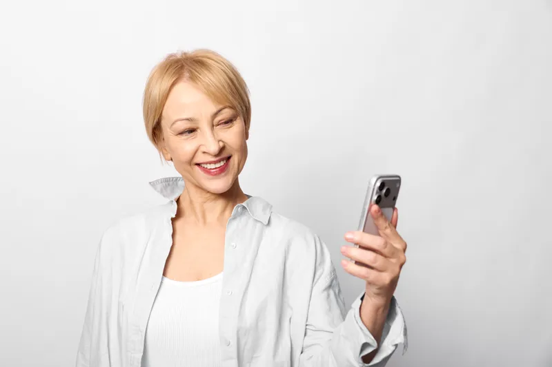 Senior woman, smiling, holding smartphone, studio portrait on white background, soft light and calm mood, communication and technology, casual home style