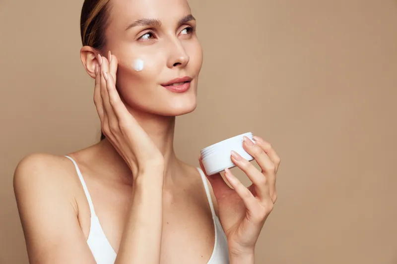 Woman applying face cream with fingers, close-up in studio light on a beige background. Skincare and self-care concept, beauty moisturizer treatment, calm wellness routine.