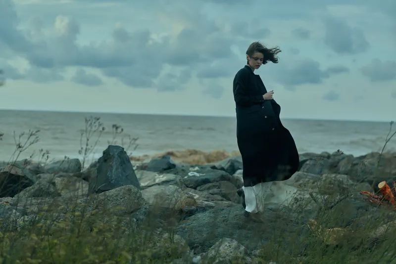 Windy coastal scene with a woman in a long coat standing on jagged rocks beside the sea, fabric fluttering in the wind, moody atmosphere, and a rugged landscape under cloudy skies.