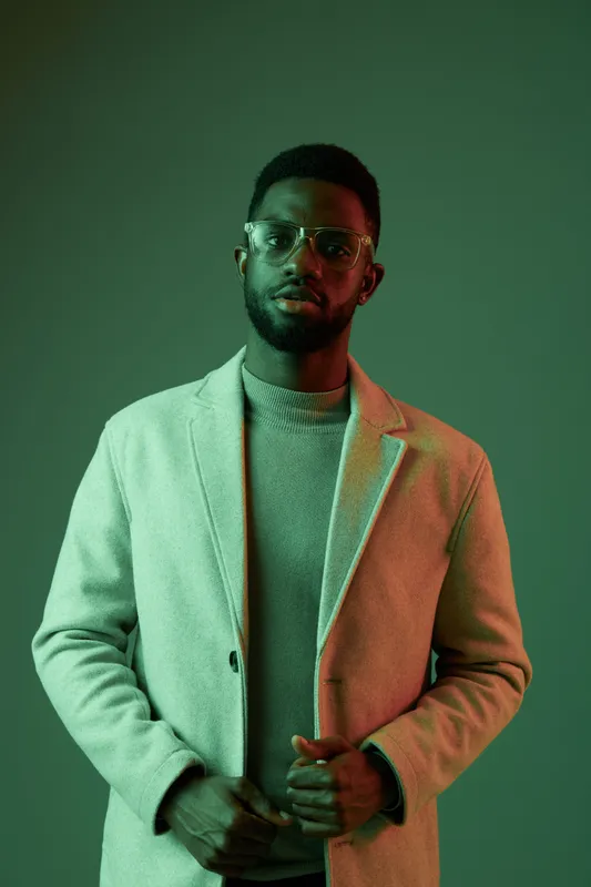 Stylish young Black man in a light coat, showcasing a confident look against a green background, reflecting modern fashion and seasonal trends