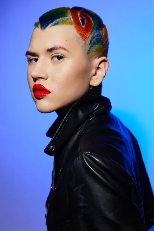 Woman with colorful rainbow hair and bold red lipstick wearing a black leather jacket, posing confidently against a vibrant blue studio background.