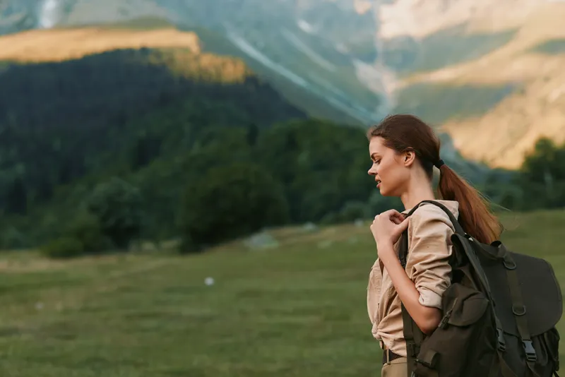 Woman hiking outdoors with backpack, looking ahead, natural light, lush landscape, distant mountains, calm moment, casual clothing, adventure vibe