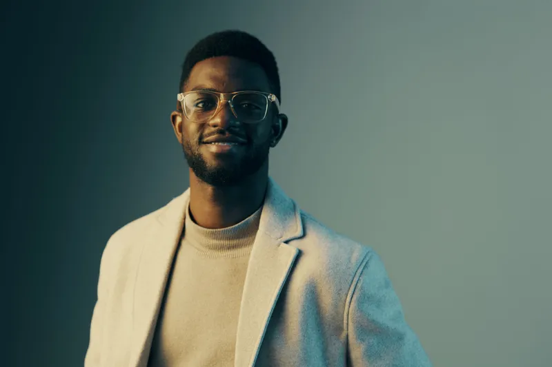 Confident Black man in stylish clothing smiling against a gradient grey background, showcasing modern fashion and personality with soft lighting