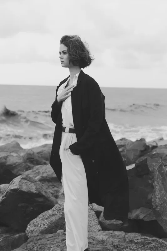 Woman standing on rocky shoreline by the sea in a long coat, wind in her hair, moody monochrome styling that highlights coastal fashion, solitude and resilience against the elements