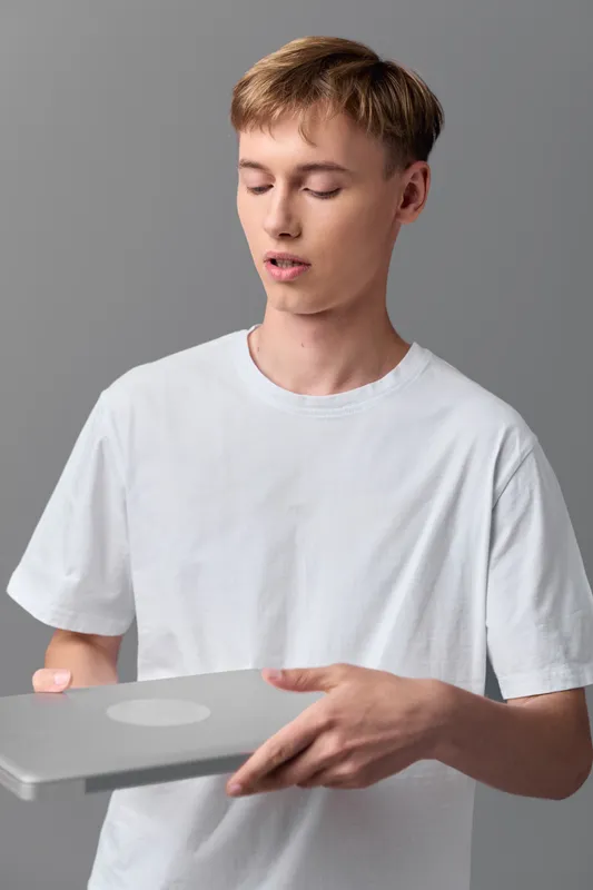 Young man in a white t-shirt holds a closed laptop in a neutral studio, emphasizing technology use, focus, and calm concentration during a casual work or study moment