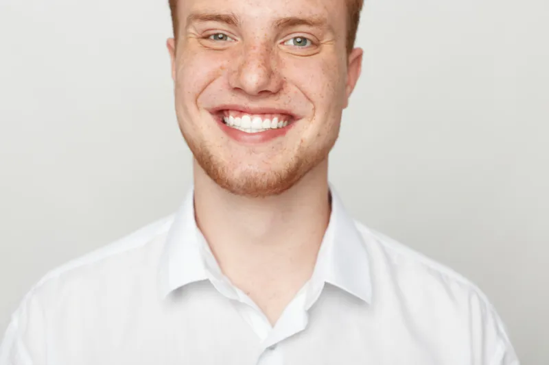 Close-up of a smiling young man with red hair and freckles wearing a white shirt. Bright studio lighting highlights his cheerful expression and natural skin texture. Positive mood.