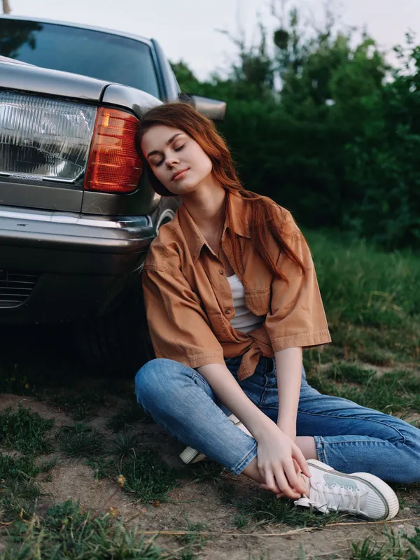 Young female model relaxing next to a vintage car, dressed in casual summer clothing, enjoying a serene moment in nature with a joyful smile.