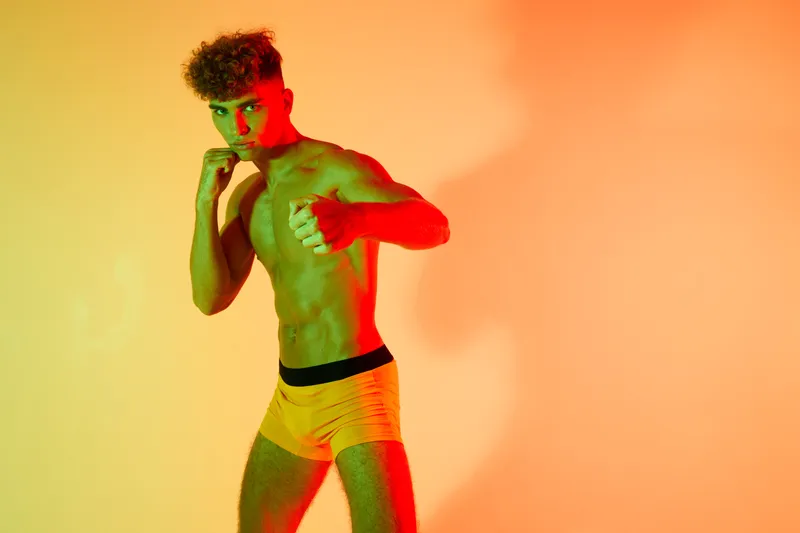 Young man with curly hair posing in fighting stance wearing yellow shorts against orange background with dramatic lighting and shadow for fitness and boxing concept.