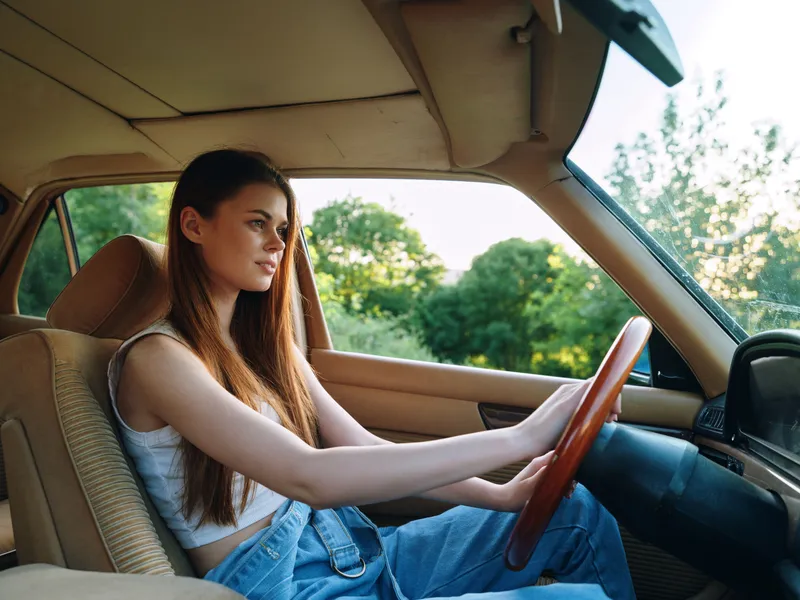 A young woman driving a vintage car surrounded by lush greenery, showcasing relaxed confidence and style on a sunny day.