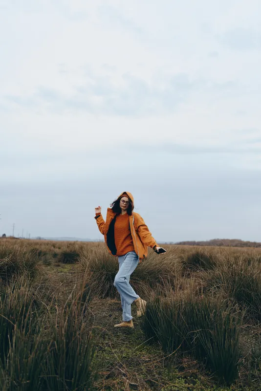 Woman outdoors lifestyle, walking through grass field, casual jeans and jacket, hands near face, cloudy sky, calm expression, natural colors, wide landscape