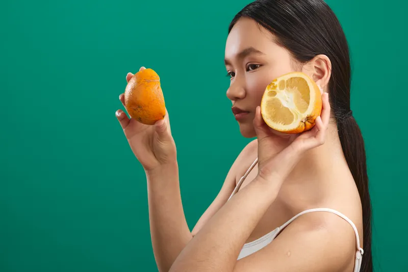 asian woman holds citrus fruits to her ears in a playful pose, contrasting bright fruit with a bold green backdrop to create a vibrant beauty moment.