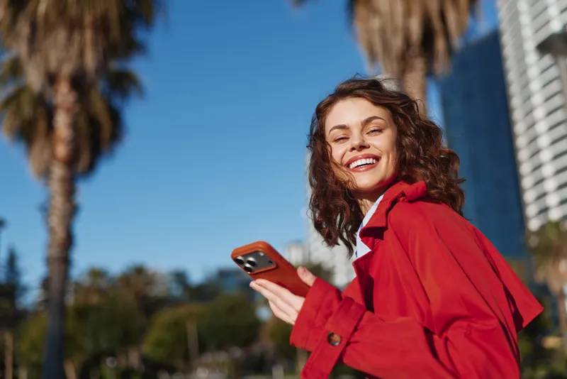 Young woman in a bright red jacket smiles while holding a smartphone outdoors in a sunny urban setting, palm trees behind, clear blue sky, casual pose and carefree vibe.