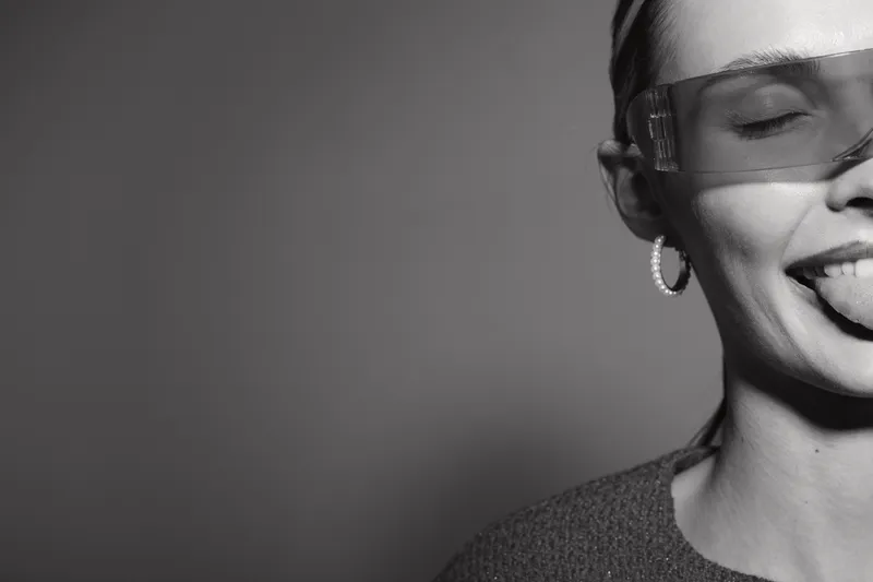 Smiling white woman with light skin and closed eyes wears transparent glasses and silver earrings, sticking out her tongue in studio. Concept of joy and playful mood.