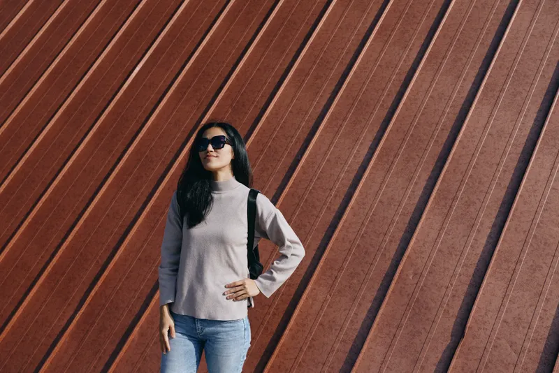 Woman wearing sunglasses and casual sweater with jeans stands against a tall striped wooden wall, showcasing street fashion, confidence, and urban lifestyle for fashion and travel.