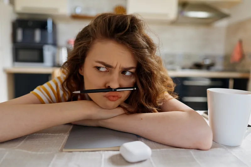 Contemplative woman in a kitchen rests her chin on a table with a pen in her mouth. Close-up reveals a notebook, laptop, mug and cluttered counter, conveying home lifestyle, daily routine, and warm,