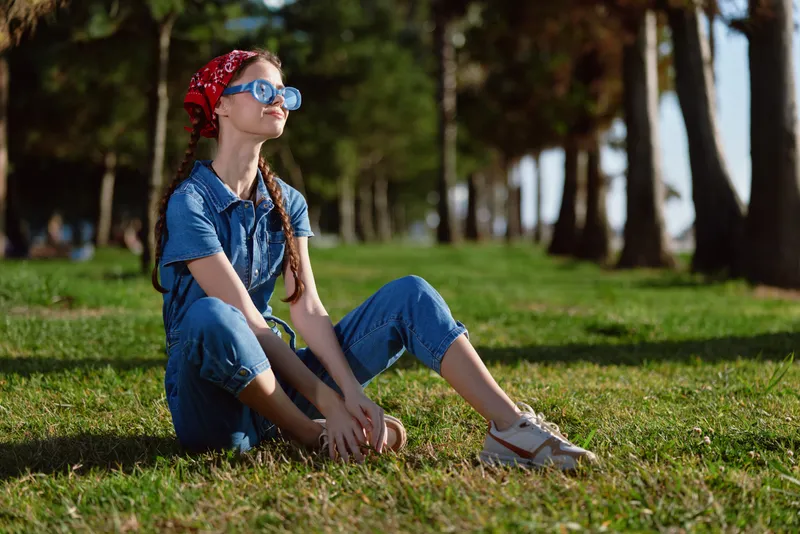 Lifestyle woman in modern boho-western denim street style sitting on grass, wearing sunglasses and a red bandana, captured with warm film color tones in the park sunlight.