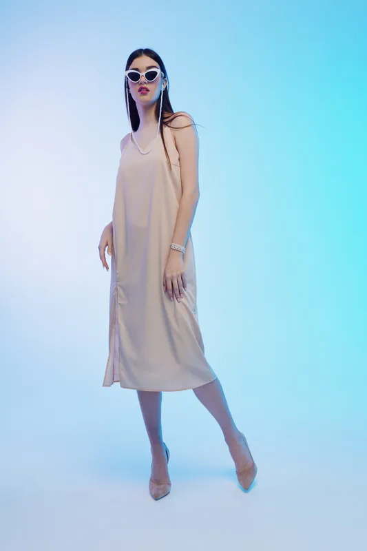 Slim woman in a beige slip dress and white sunglasses poses confidently on a blue gradient background. Concept of fashion, beauty, and modern style in studio lighting.