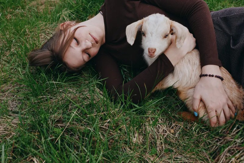 Lifestyle moment with a woman outdoors, lying on grass and cuddling a young goat, peaceful bond, gentle touch, natural colors, serene mood, candid and warm light in a rural setting