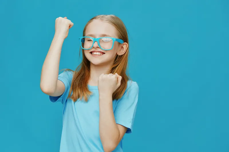 Happy girl celebrating success with colorful glasses, dressed in a light blue t shirt, against a vibrant turquoise background, embodying joy and positivity