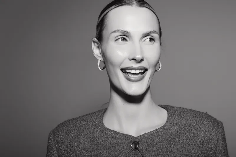 Smiling white woman with fair skin and light hair wears textured sweater and earrings against simple background. Portrait captures joyful expression and natural beauty concept.