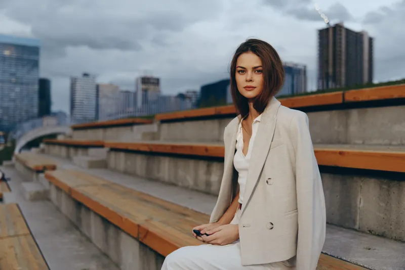 Confident business woman in beige suit sitting outdoors on urban benches with city skyline background, modern professional, autumn corporate style