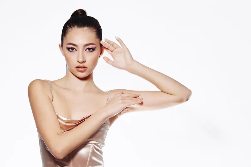Elegant young woman wearing a satin dress posing with hand gestures near her face against a white background. Creative beauty and fashion concept highlighting style and confidence.