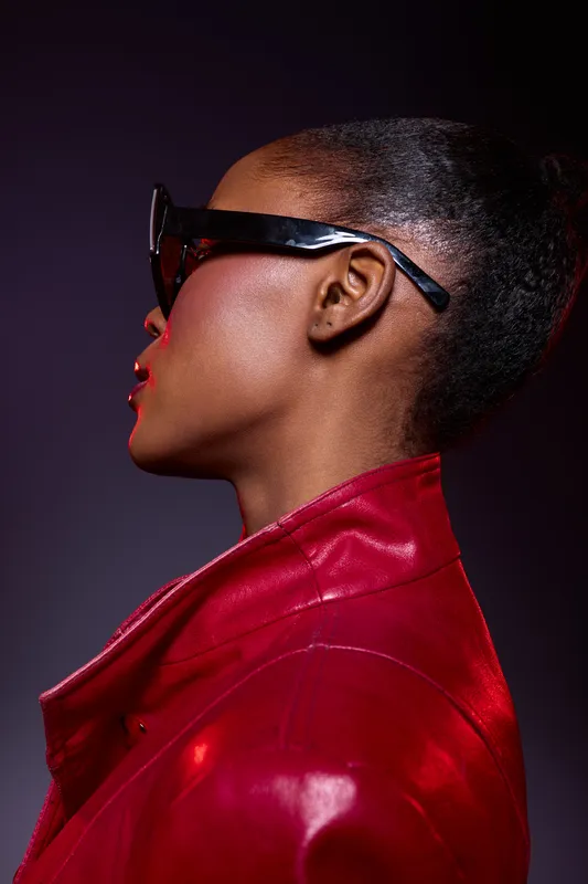 Profile of a young black woman wearing black glasses and a red leather jacket. The studio portrait highlights her dark skin and confident pose against a dark background.