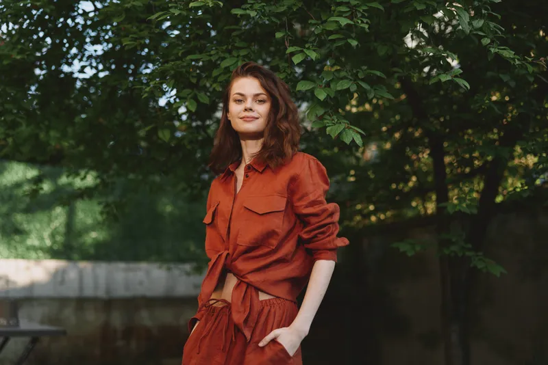 Smile on a woman in a rust dress posing outdoors in a park, surrounded by greenery. Natural light highlights casual fashion and confident, relaxed lifestyle, capturing everyday elegance and warmth