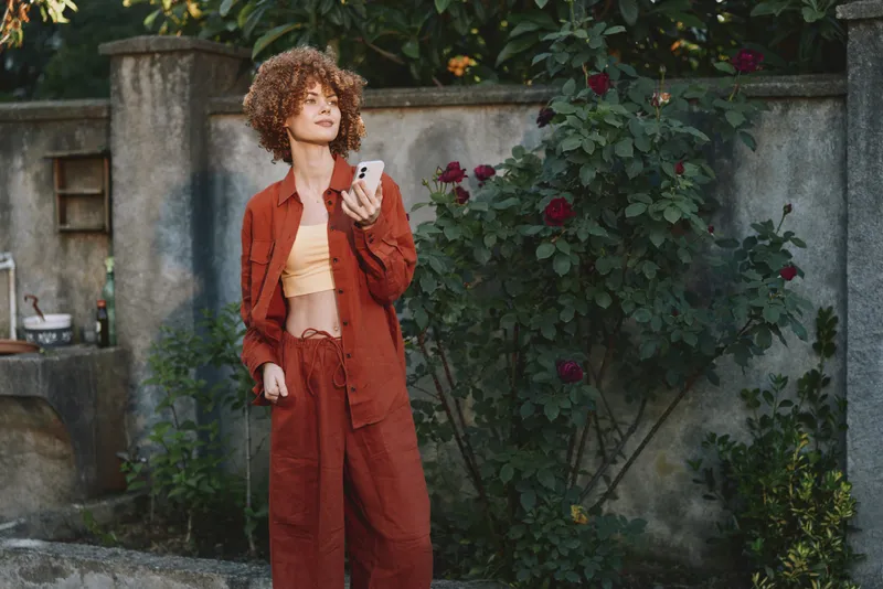 Curly woman in a rust suit stands in a lush garden, looking at her smartphone with a confident, relaxed expression, daylight framing the scene and potted plants around her, conveying casual chic