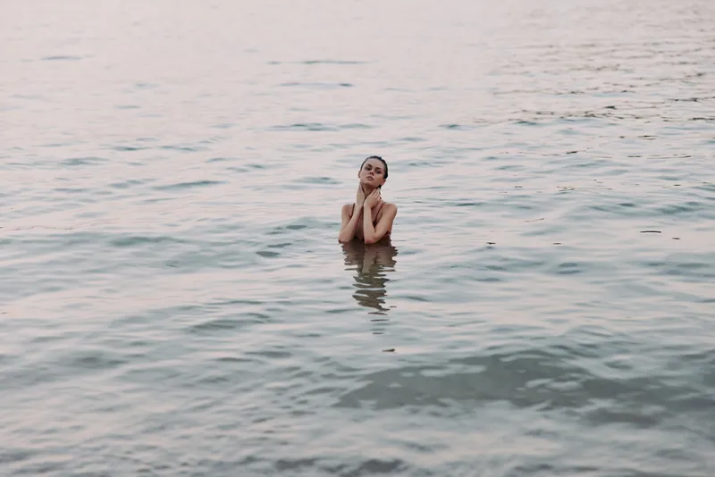 A lone figure stands waist deep in calm sea water, hands near face, conveying introspection as soft light glints on the rippling surface.