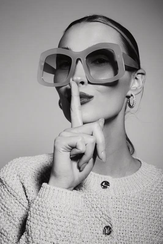 Stylish caucasian woman with fair skin wearing large sunglasses makes silence gesture with finger on lips.