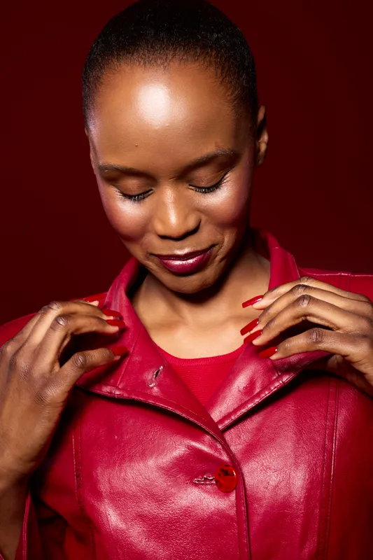Portrait of elegant black woman with dark skin wearing red leather jacket, looking down and adjusting collar. Studio shot with burgundy background. Concept of fashion, confidence, and style.