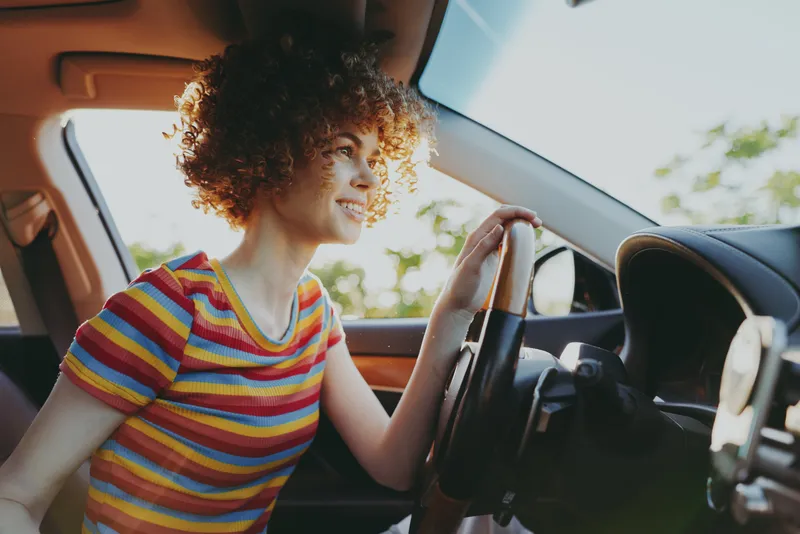 Smiling woman with curly hair driving a car, wearing a striped shirt. Bright colors and warm light create a retro film mood that highlights everyday travel and casual lifestyle.