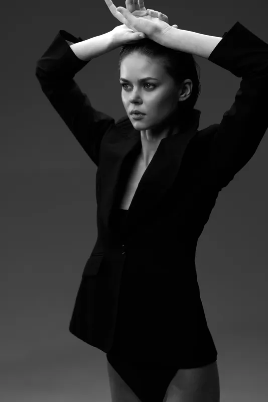 Black and white portrait of a serious woman wearing a blazer with arms raised in studio lighting.