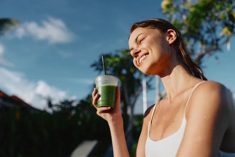 A woman outdoors drinks a green smoothie, smiling under bright sun, showcasing a healthy lifestyle moment. Focus on wellness and refreshment in a casual, sunlit backyard setting.