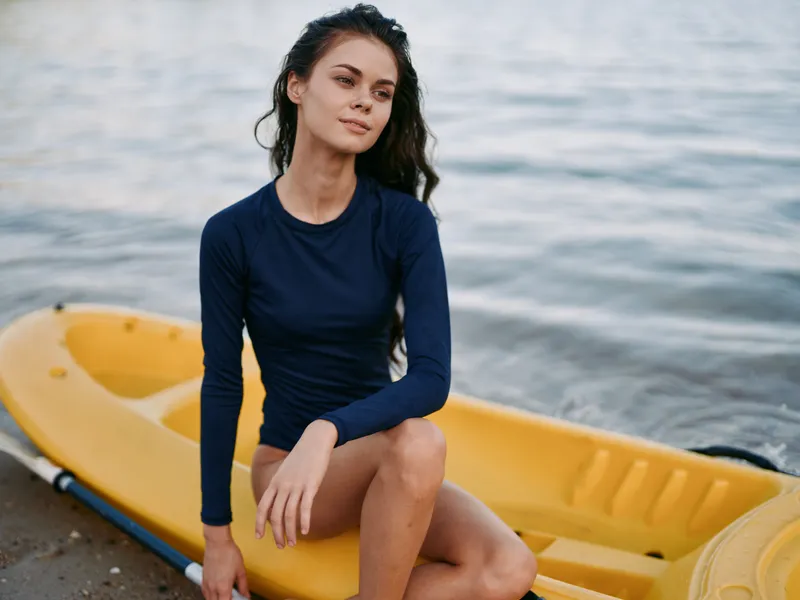 Young woman sits beside a yellow kayak on a calm shoreline, wearing a navy long sleeve top. Tranquil glow highlights a confident pose by open water.
