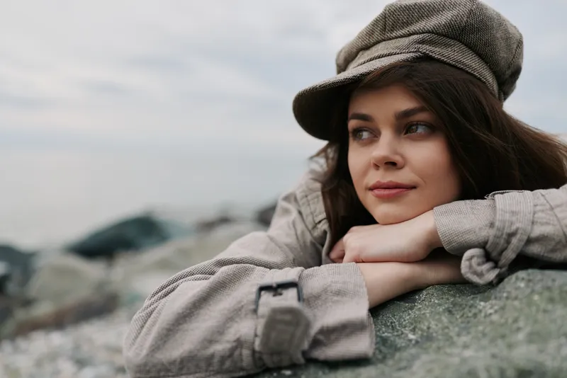 Young woman in casual jacket and cap leans on rock outdoors, looking away thoughtfully. Overcast sky and blurred rocky background create a calm, reflective mood in nature.
