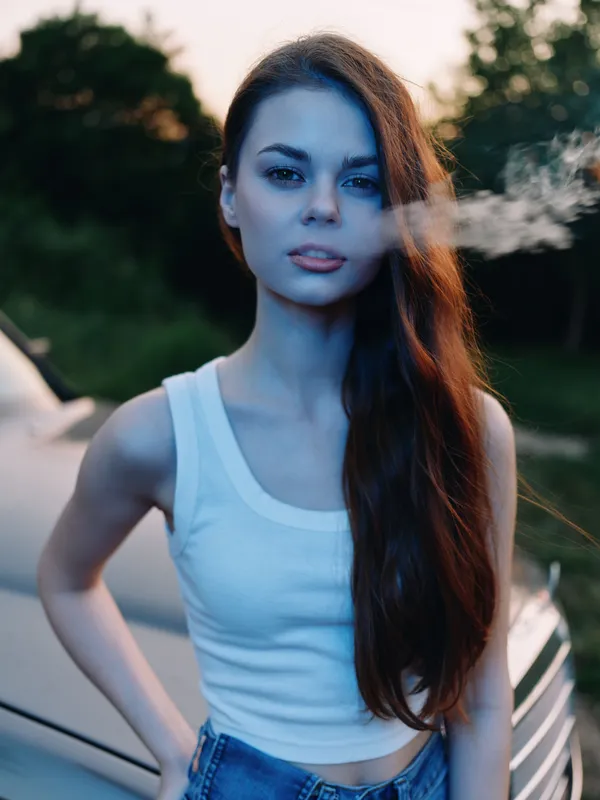 Young woman exhaling smoke in a serene outdoor setting, wearing a white tank top and denim shorts, embodying a sense of freedom and carefree spirit.