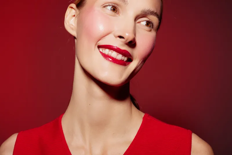 Smiling woman with red lipstick and red dress looks away on red background. Close-up portrait with bright makeup and smooth skin. Concept of beauty, fashion, and confidence.