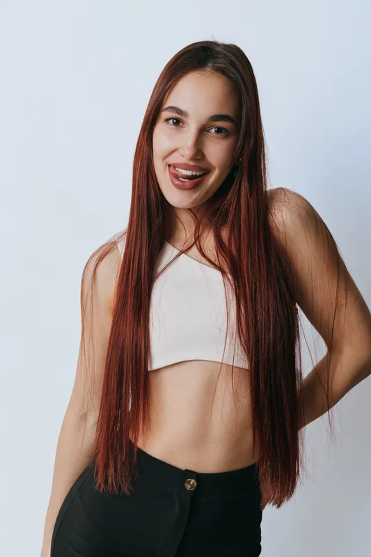 Young woman with long straight hair smiling and posing in casual outfit with white crop top and black pants on light background. Attractive female model with natural makeup and confident expression.