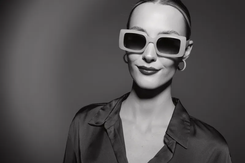 Elegant white woman with fair skin wearing large sunglasses and silky blouse smiles confidently on neutral background. Concept of fashion, style, portrait, and modern accessories.