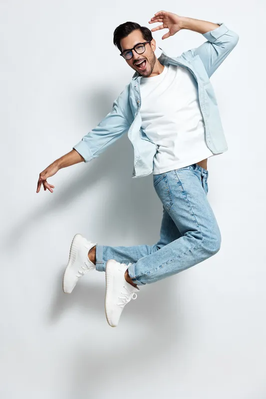 Full body smiling cheerful young man wearing casual outfit poses in playful jump, expressing joy and energy against a soft gray background. Lifestyle concept